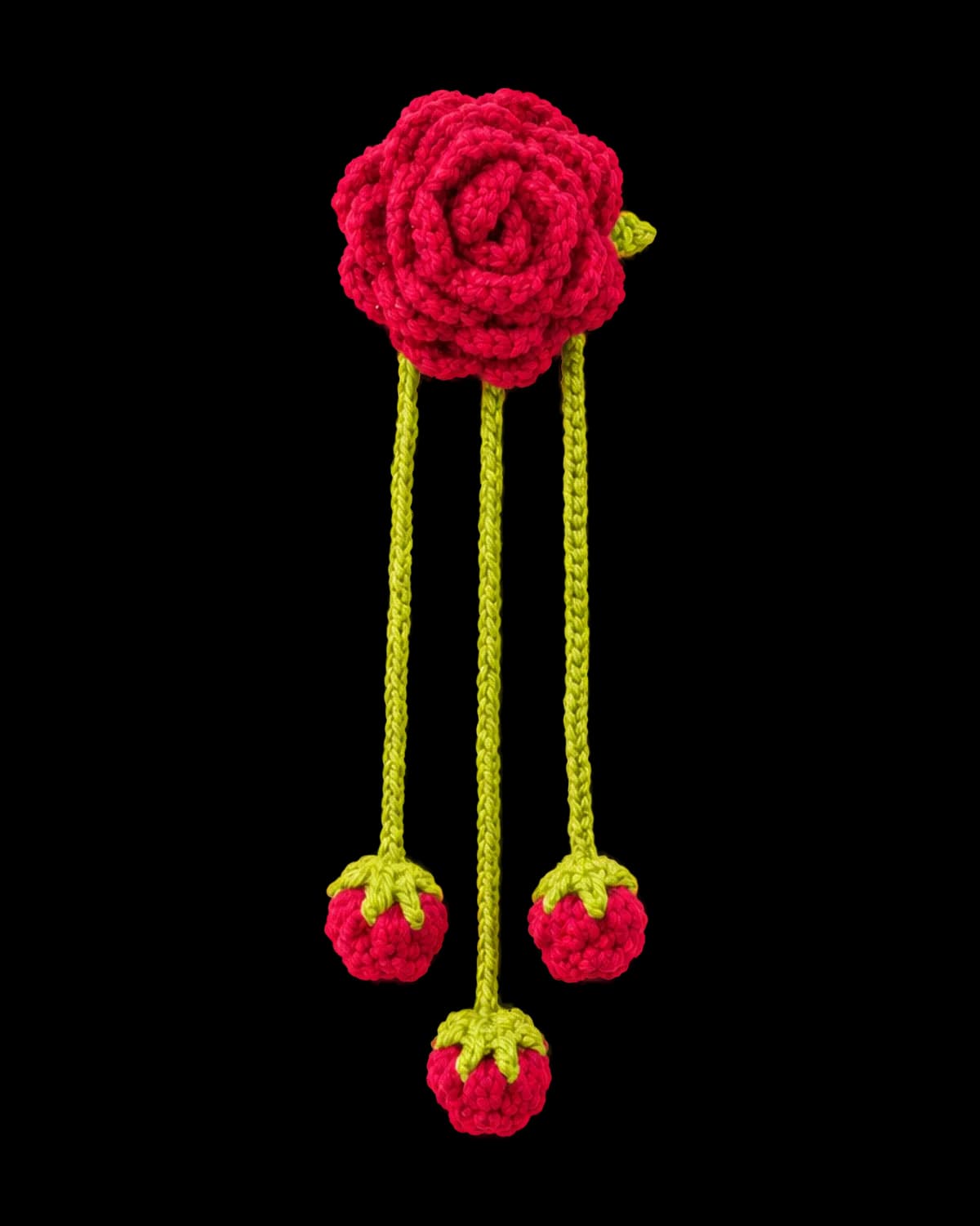 Crochet hair clip with a deep raspberry-red rose, a small green leaf and three lime-green cords ending in red crochet strawberries, on a cream background